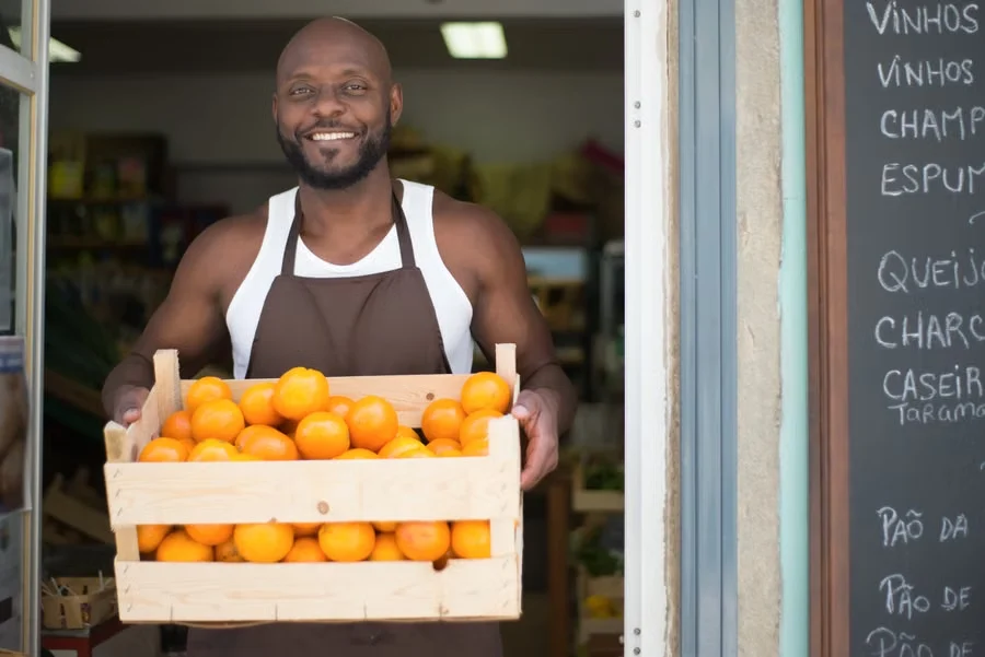 A shop owner in an apron carrying a crate of oranges in the doorway